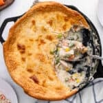 A skillet chicken pot pie with a golden, flaky crust, partially served to reveal its creamy Skillet Chicken Pot Pie filling of chicken, peas, carrots, corn, and green beans. A serving spoon rests inside the dish.