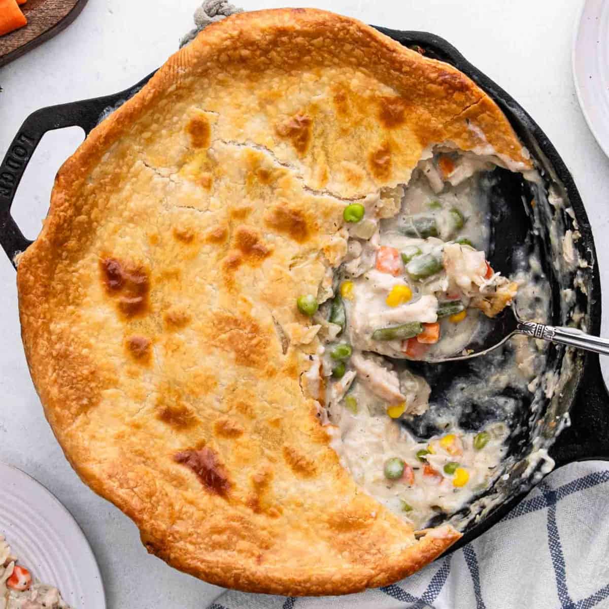 A skillet chicken pot pie with a golden, flaky crust, partially served to reveal its creamy Skillet Chicken Pot Pie filling of chicken, peas, carrots, corn, and green beans. A serving spoon rests inside the dish.
