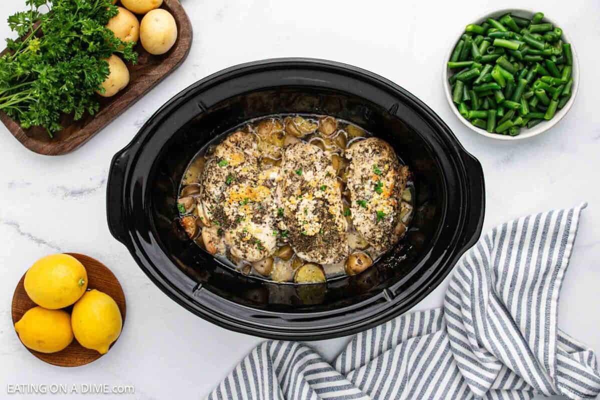 Cooked chicken and potatoes in the crock pot with a small bowl of green beans, fresh lemon and small potatoes on the side.