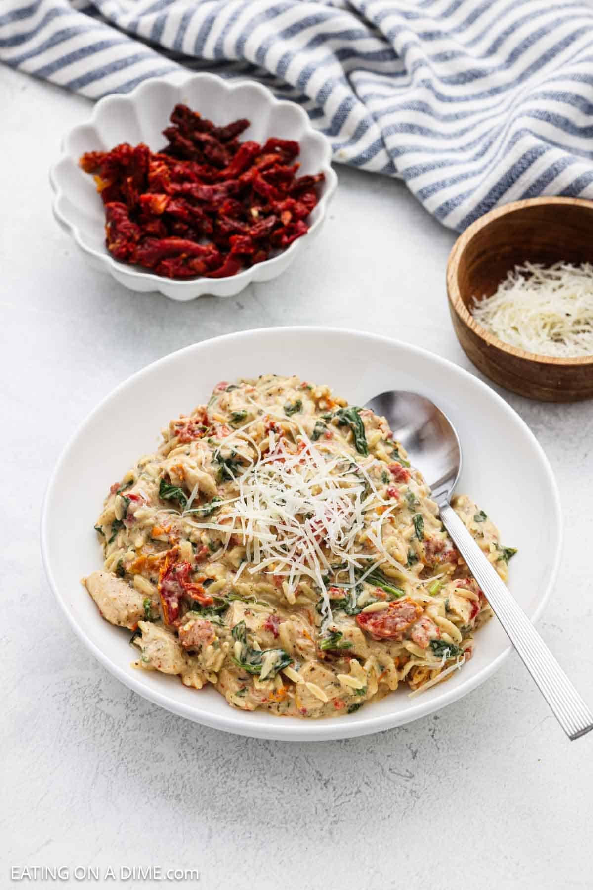 A serving of the chicken orzo in a white bowl topped with shredded parmesan cheese and small bowls of sun dried tomatoes and parmesan cheese on the side.