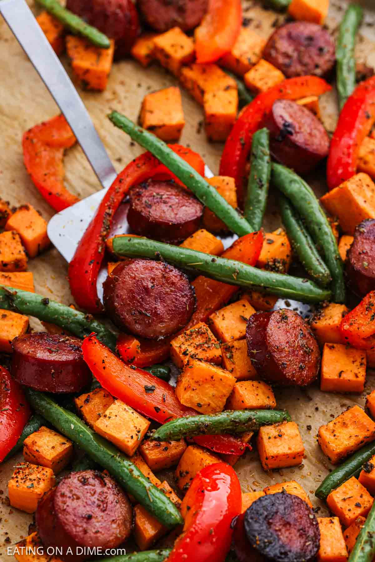 Roasted slice sausage and veggies on a sheet pan with a serving on a spatula.