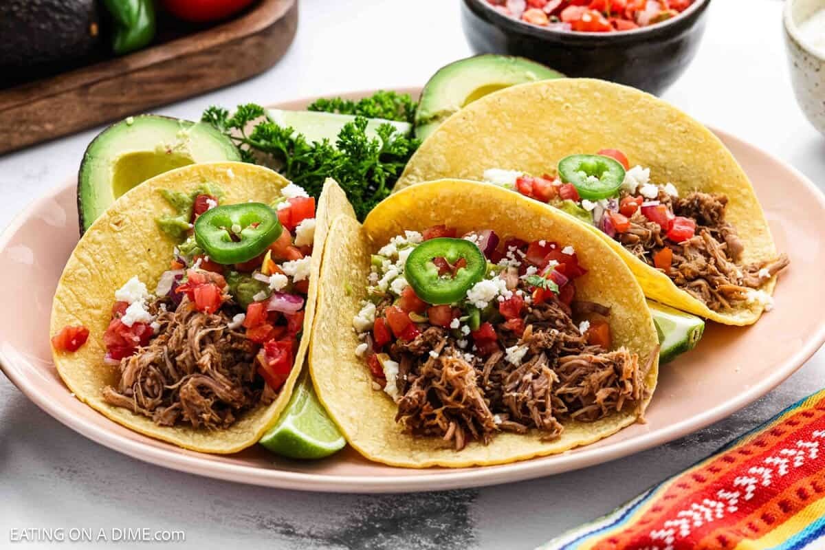 3 Pulled Pork Tacos on a plate topped with sliced jalapenos, pico de gallo, crumbled cheese with a side of avocados.