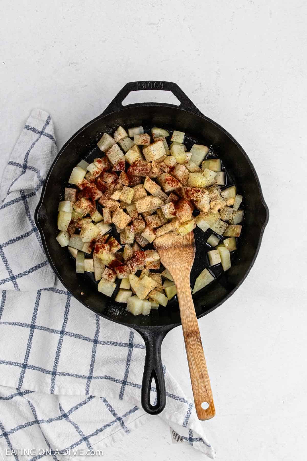 Diced potatoes in a cast iron skillet topped with spices with a wooden spoon.
