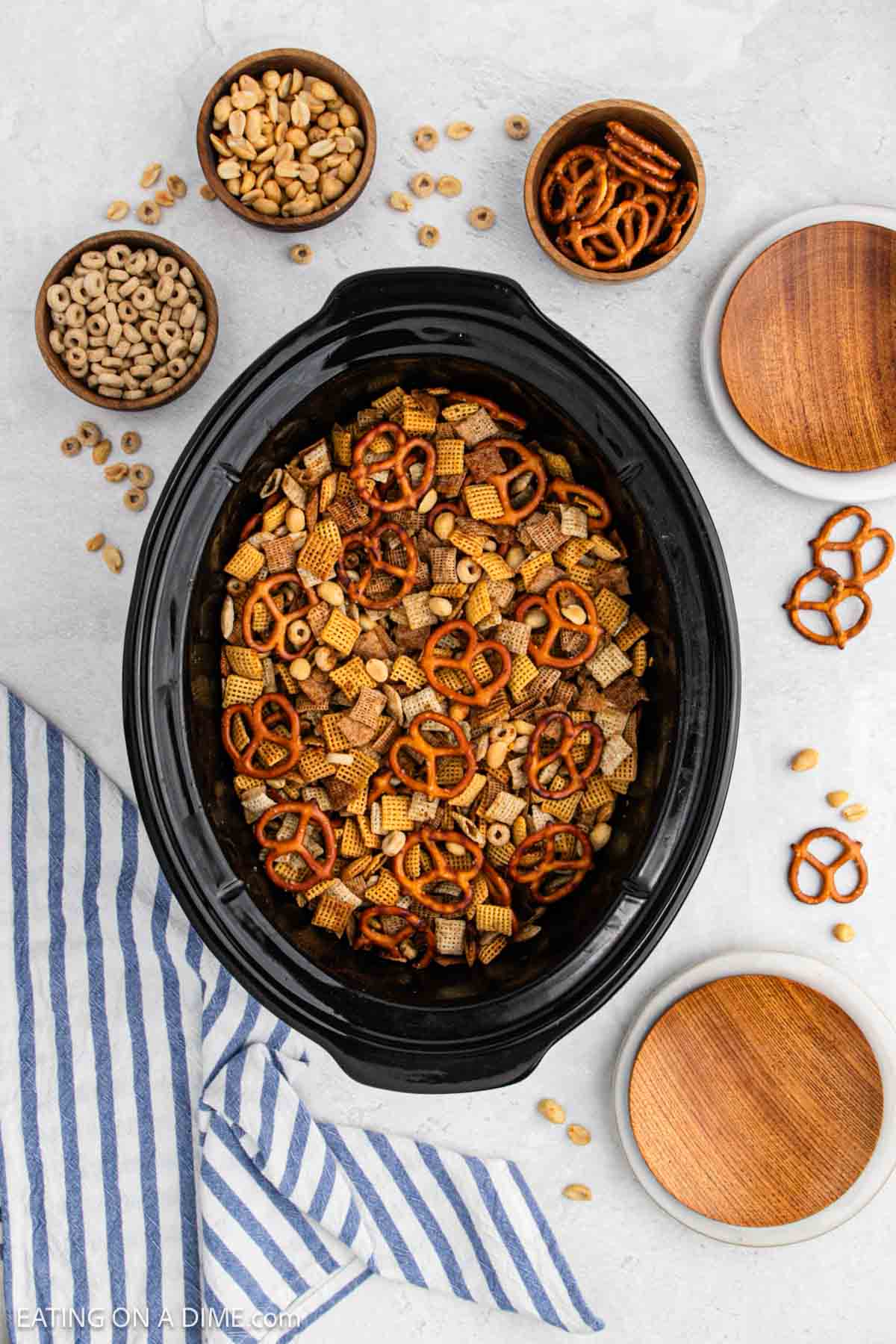 Cooked chex mix in the slow cooker with small bowls of peanuts, cheerios, and pretzels nearby.