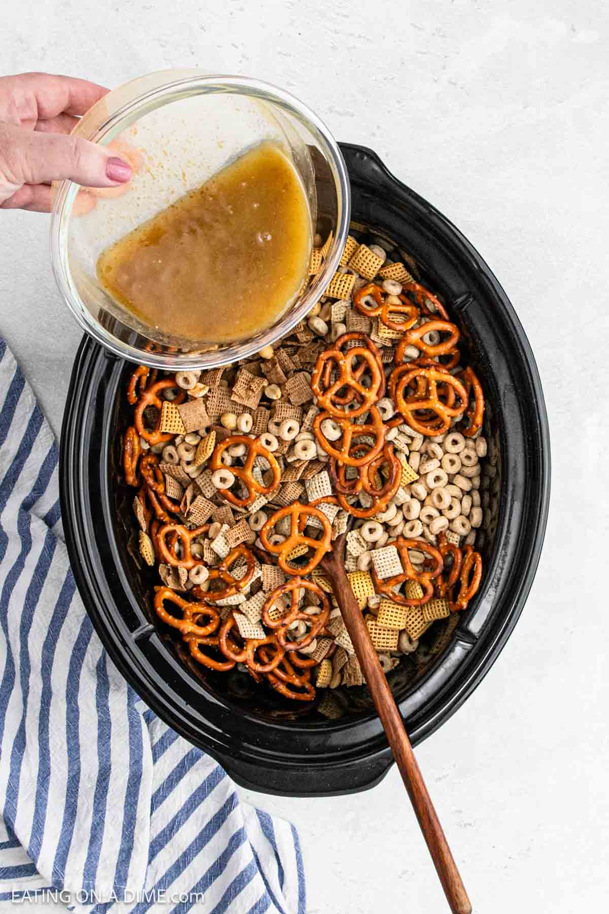 Pouring the melted butter mixture over the Chex Mix mixture in the slow cooker with a wooden spoon.