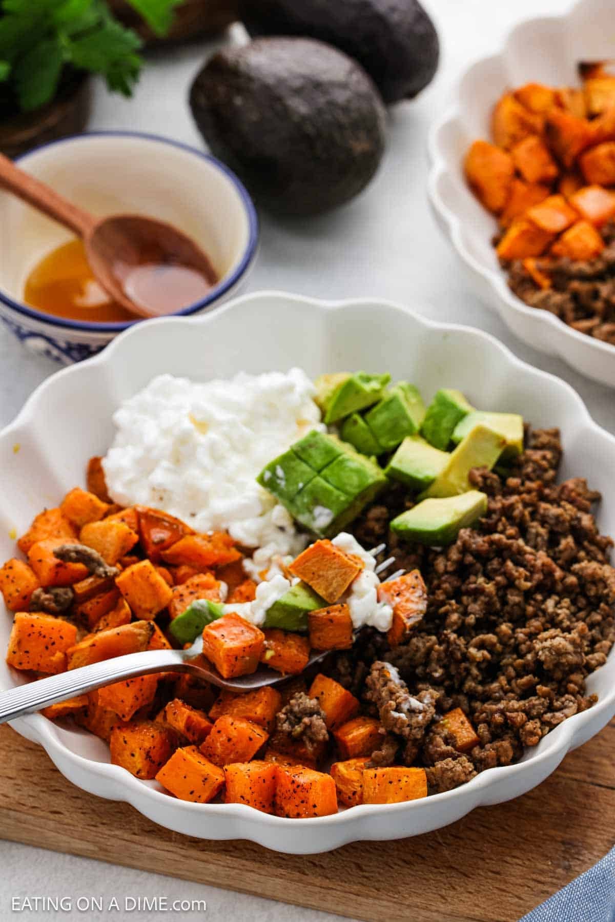 Cooked ground beef, diced sweet potatoes, cottage cheese and diced avocados in a white bowl with a fork ready to take a bite.