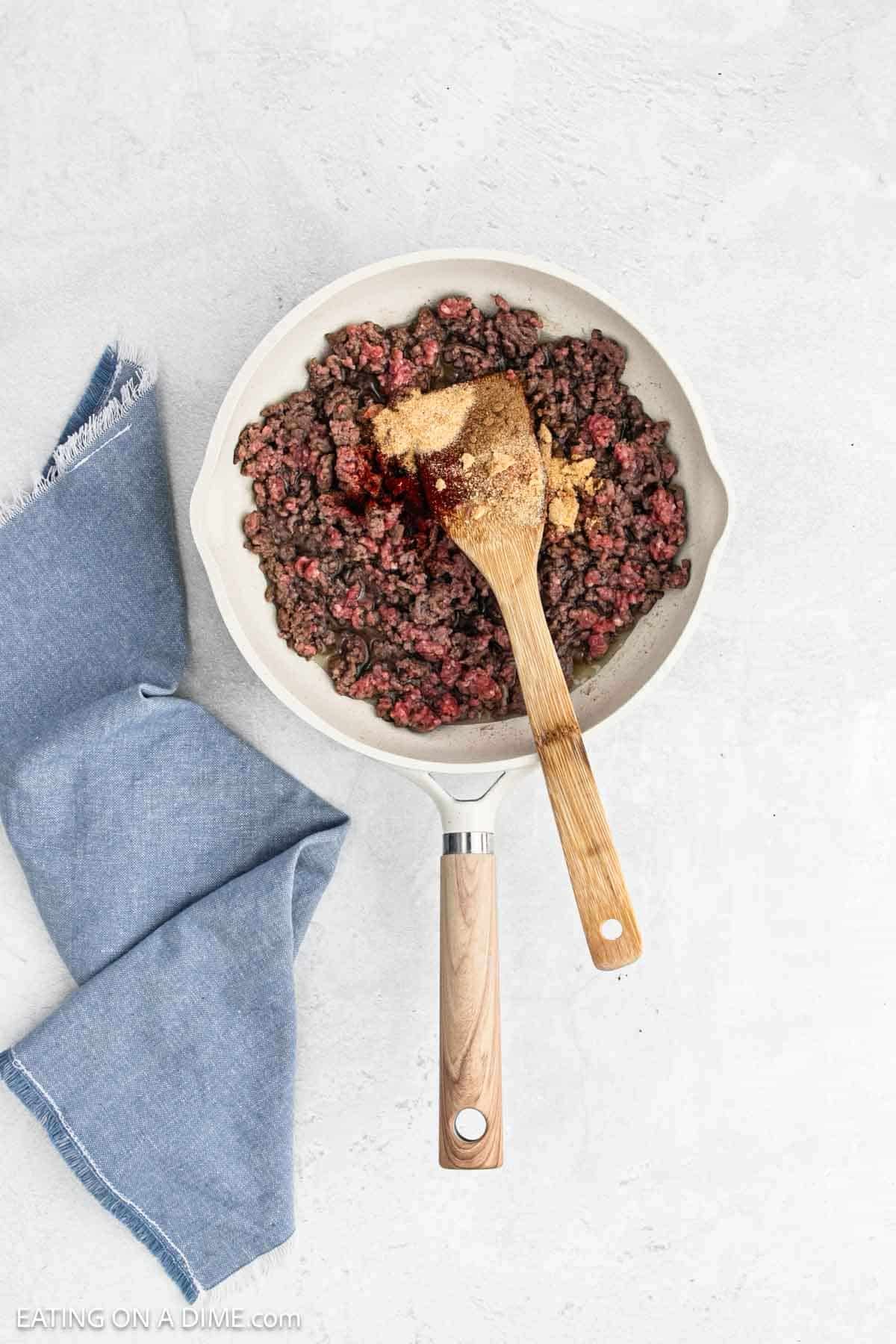Cooking ground beef in a large skillet with a wooden spoon topped with spices.