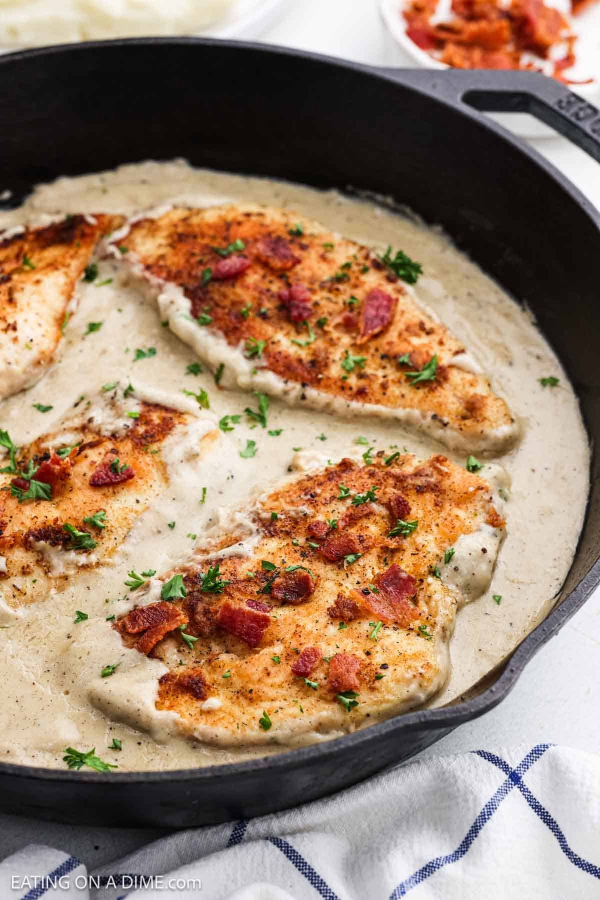 Golden brown chicken breast topped with crispy bacon submerged in gravy in a cast iron skillet.