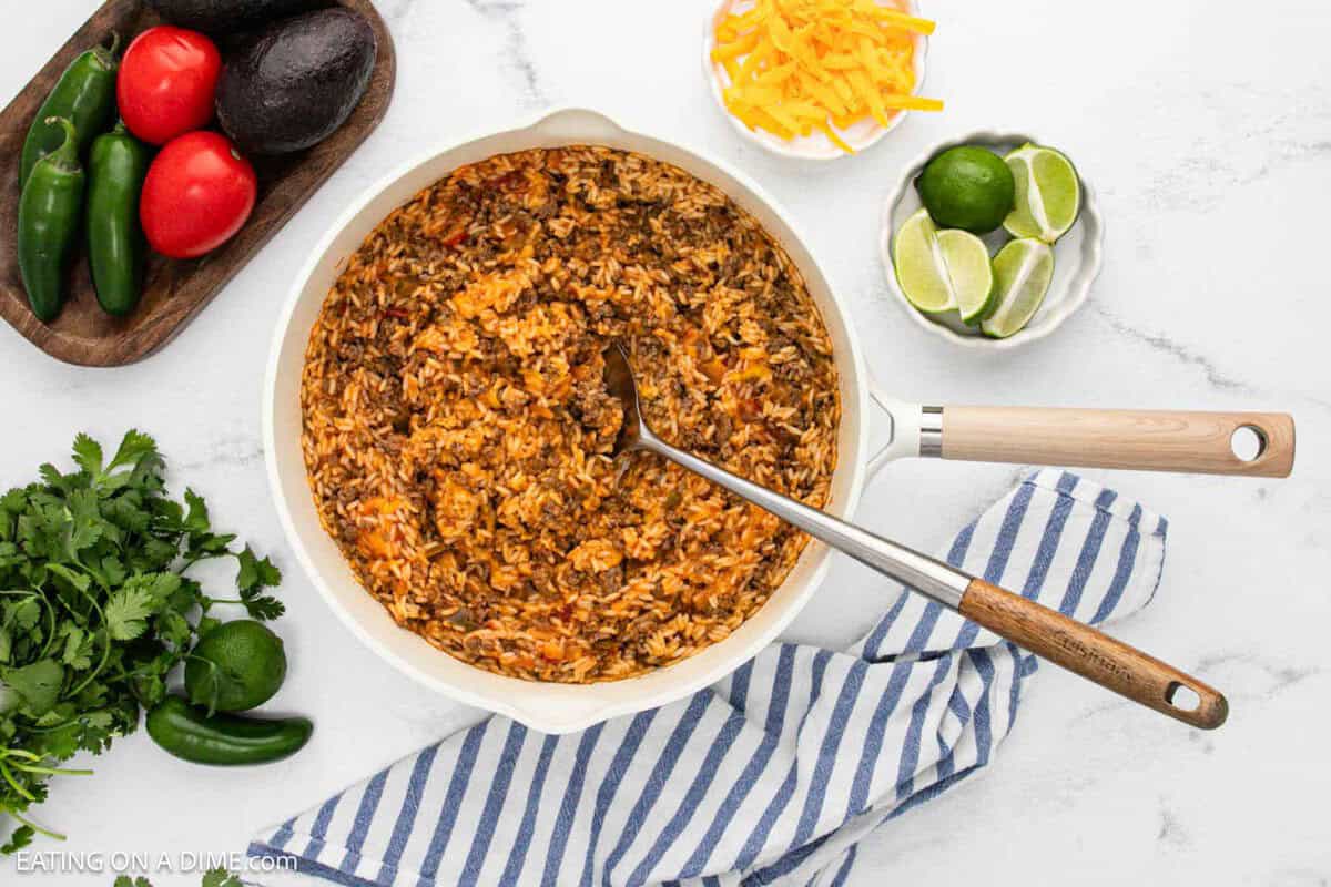 Taco rice in a large skillet with small bowls of shredded cheese and lime wedges on the side.