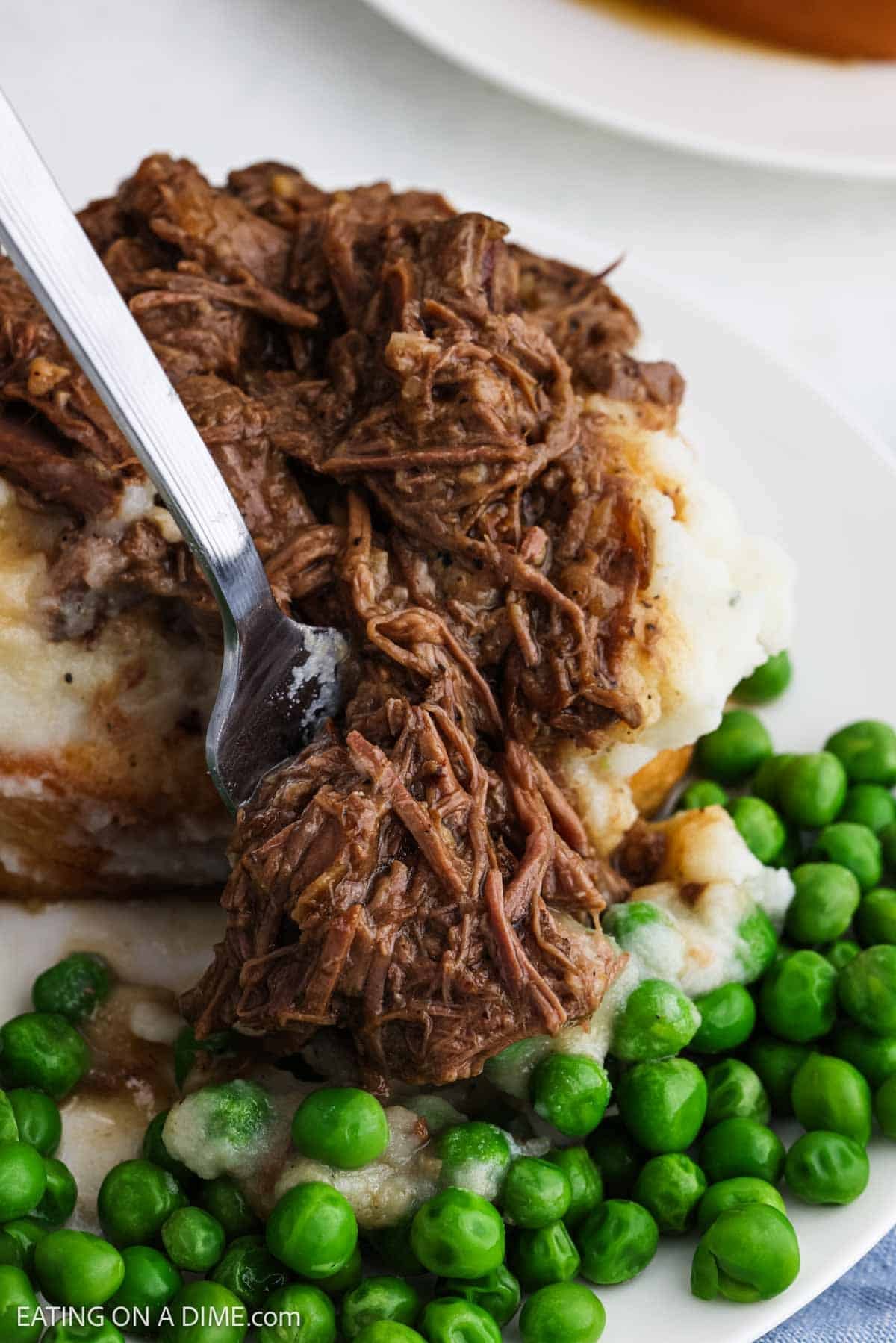 A serving of shredded beef over mashed potatoes with a side of peas.