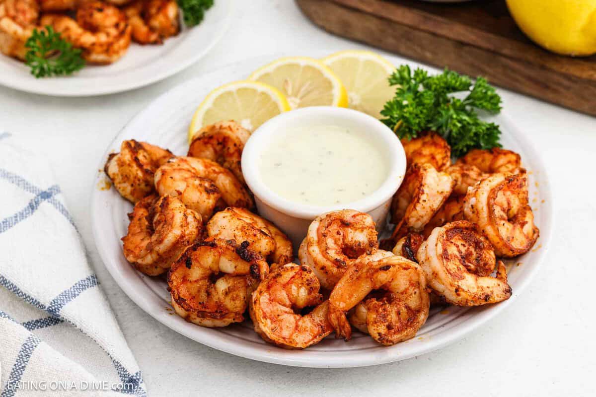 Blackened Shrimp stacked on a white plate with a bowl of sauce in the center with a side of lemon slices. 