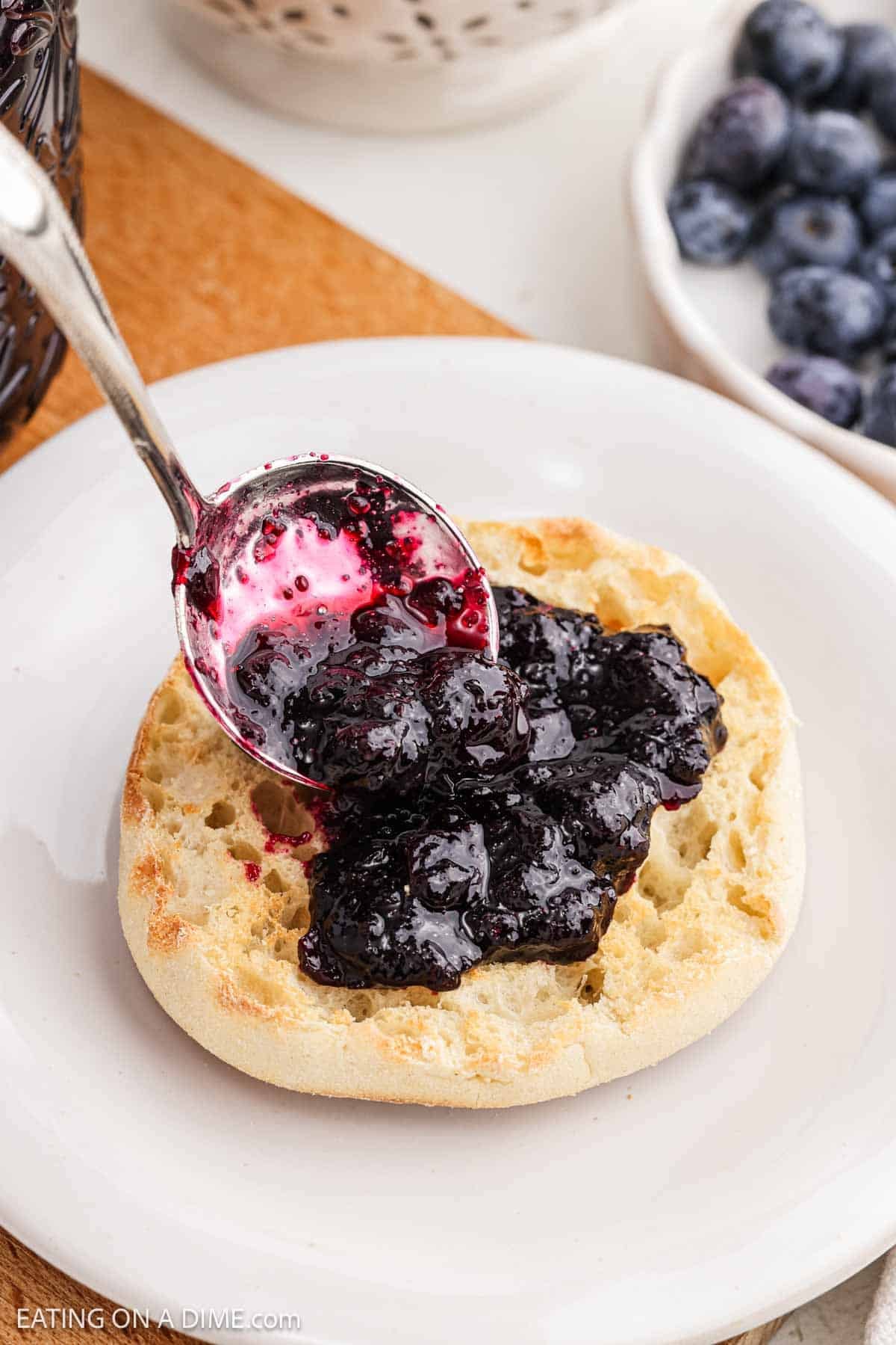 Jam being spread over a English Muffin on a plate.