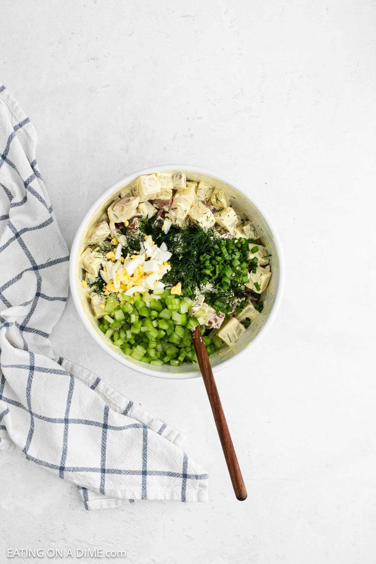 Topping the red potatoes mixture with celery, dill, and hard boiled eggs in a bowl with a wooden spoon.