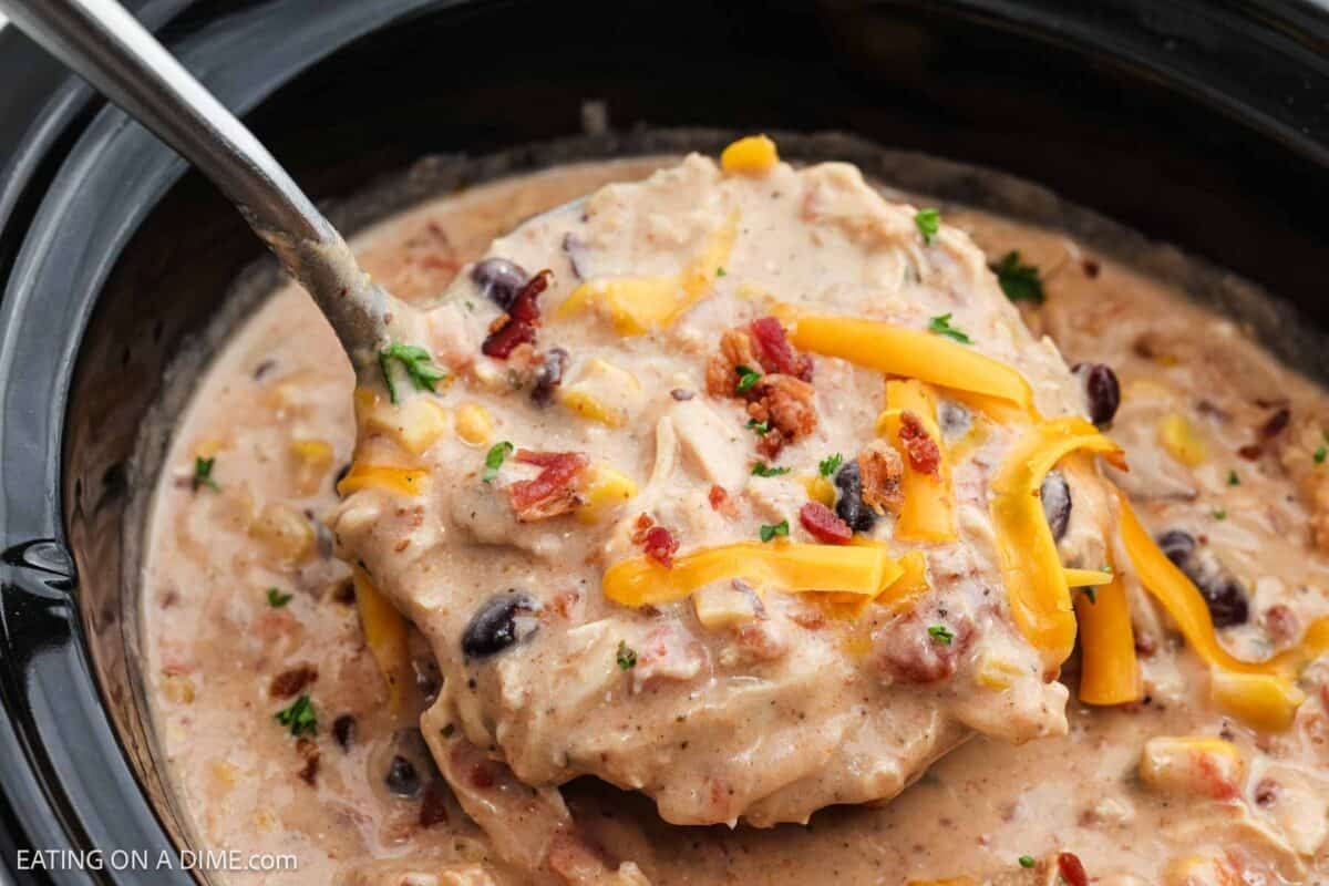 Crack chicken chili in the slow cooker topped with shredded cheese with a serving on a ladle.