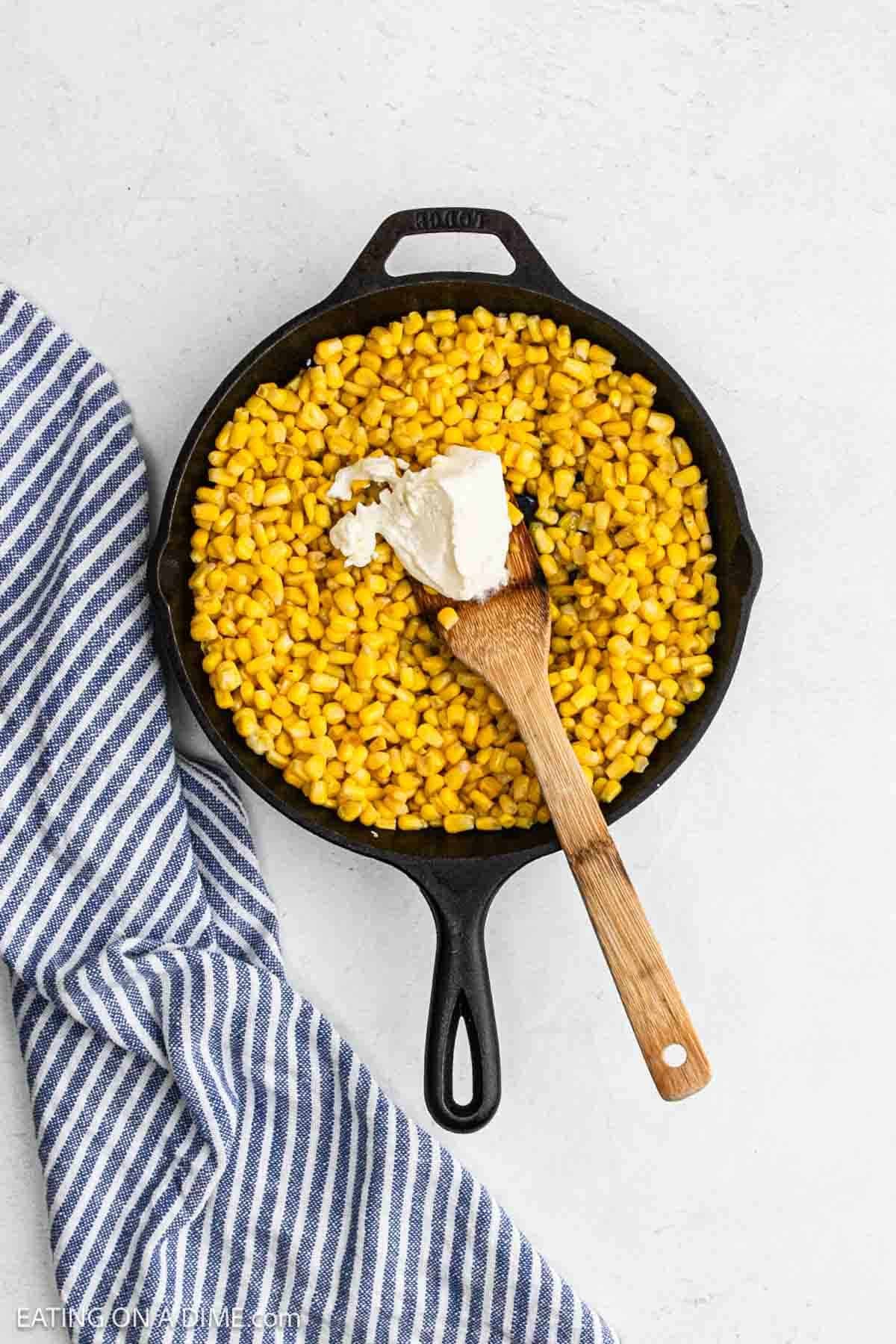 Cream cheese added to the corn kernels in a cast iron skillet with a wooden spoon.