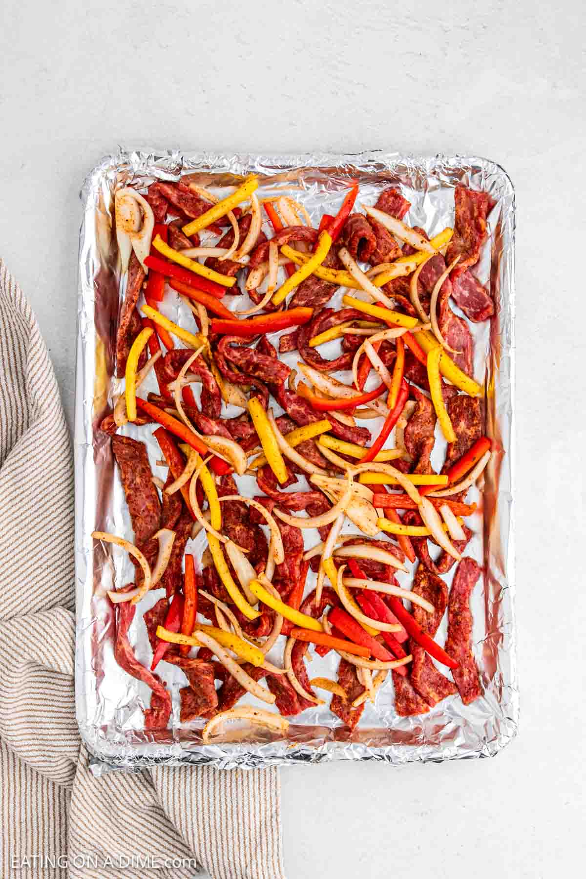 Steak strips, bell peppers and onions on a baking sheet lined with foil.